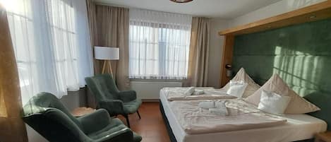 Et hotelværelse med to senge, et siddeområde med stole, en lampe og et stort vindue med gardiner.