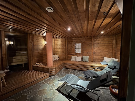 A log cabin interior with a stone floor, wooden walls, a central pillar, a window, and a seating area with chairs and cushions.