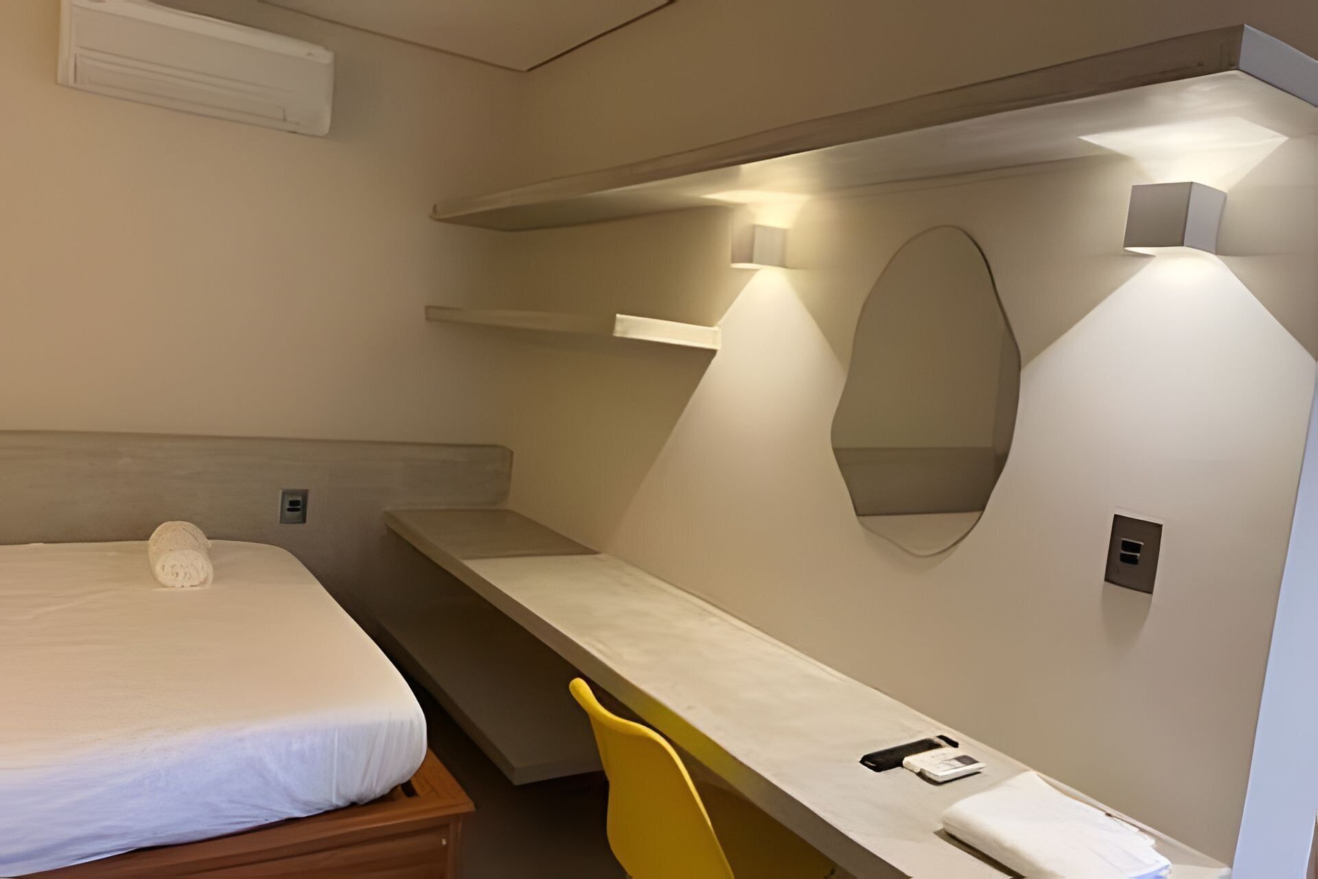 A hotel room with a bed, a desk, a chair, a mirror, and a wall-mounted light fixture.