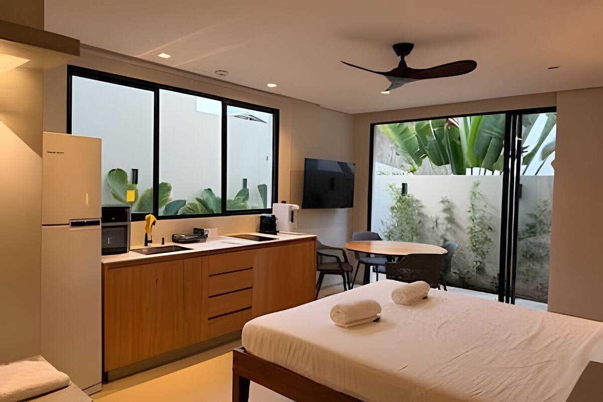 Quarto de hotel moderno com cama, escrivaninha, cadeira, pequena área de cozinha e janelas amplas.