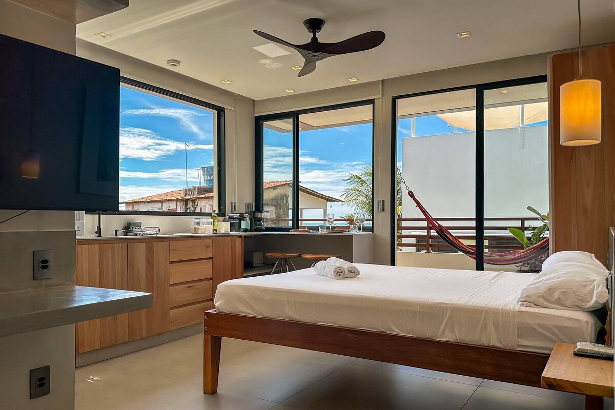 Quarto de hotel moderno com cama, televisão, varanda com rede e vista para o oceano.