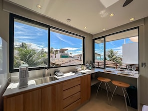 A modern kitchen with large windows, a built-in sink, and a bar area with stools. - Chalé by Vila Manga Rosa (Tibau do Sul)