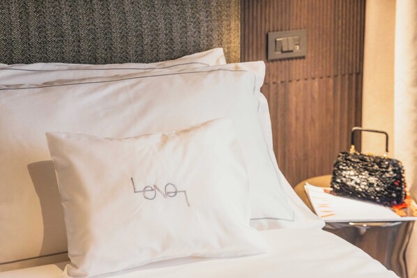A hotel room with a neatly made bed, a pillow with a name tag, a small table with a handbag, and a wall-mounted light switch. - Lenel Boutique Room - La Locanda (Brescia)