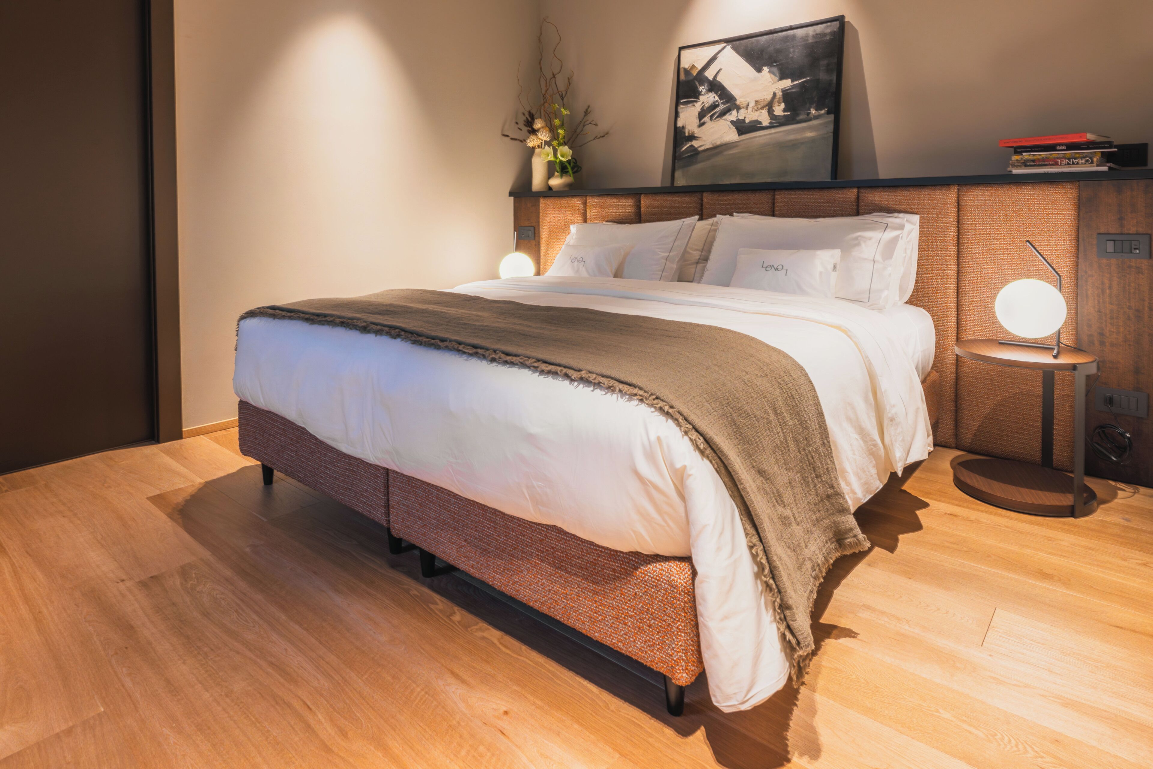 Uma cama bem arrumada com cabeceira, mesa de cabeceira com abajur e um quadro na parede.