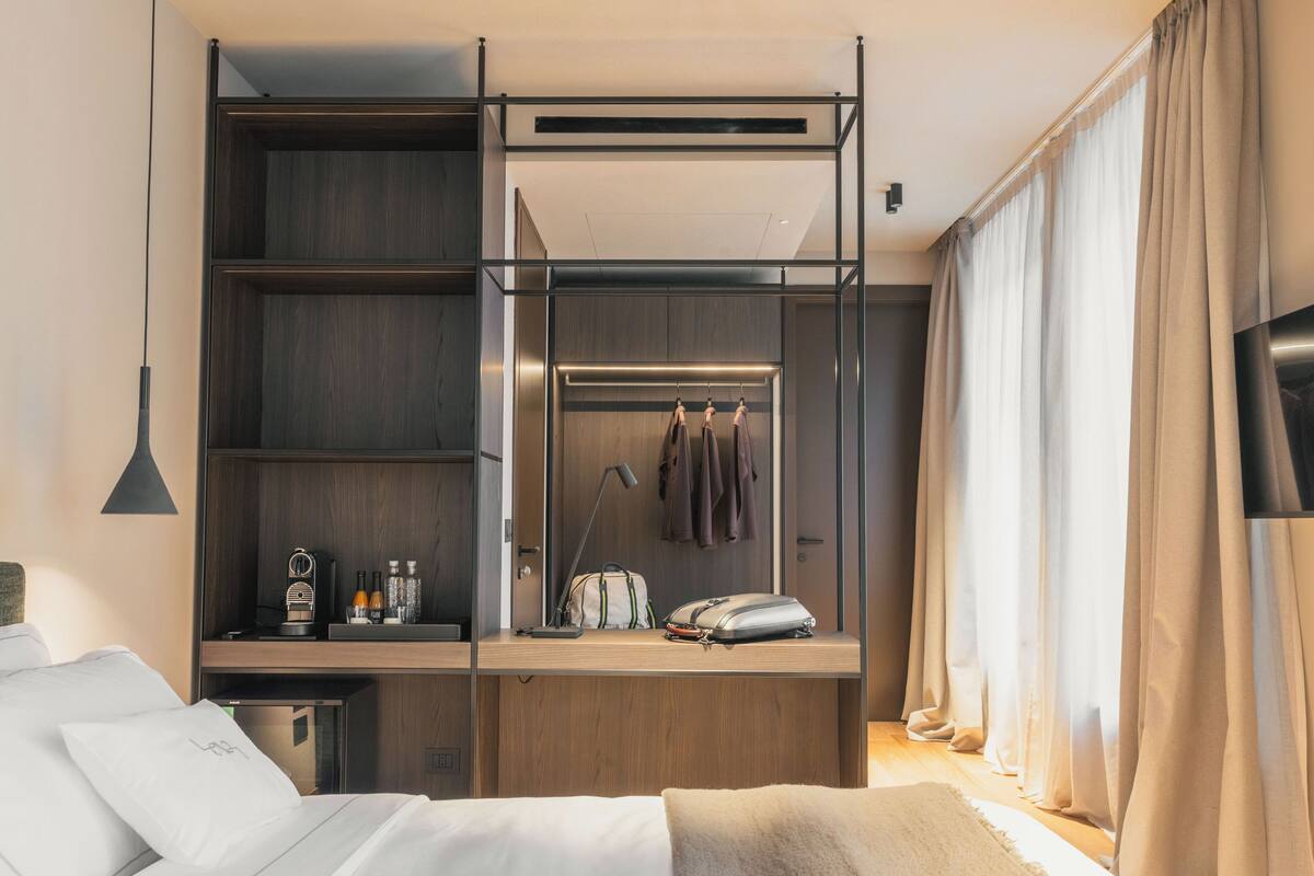 Moderni hotellihuone, jossa on sänky, työpöytä lampun kanssa, vaatekaappi, jossa on henkarit, ja seinälle asennettu televisio.