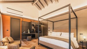 Una cama con dosel y cabecera, una zona de estar con una silla y reposapiés, un armario y una mesilla de noche con lámpara.