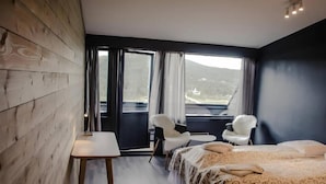 A hotel room with a wooden wall, a bed with patterned bedding, a white chair, a small table, and a large window with curtains. - Rondablikk (Nord-Fron)