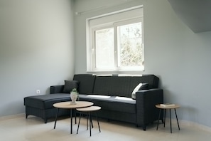 Ein modernes Wohnzimmer mit einem grauen Sofa, zwei runden Beistelltischen und einem großen Fenster mit Blick auf Bäume.