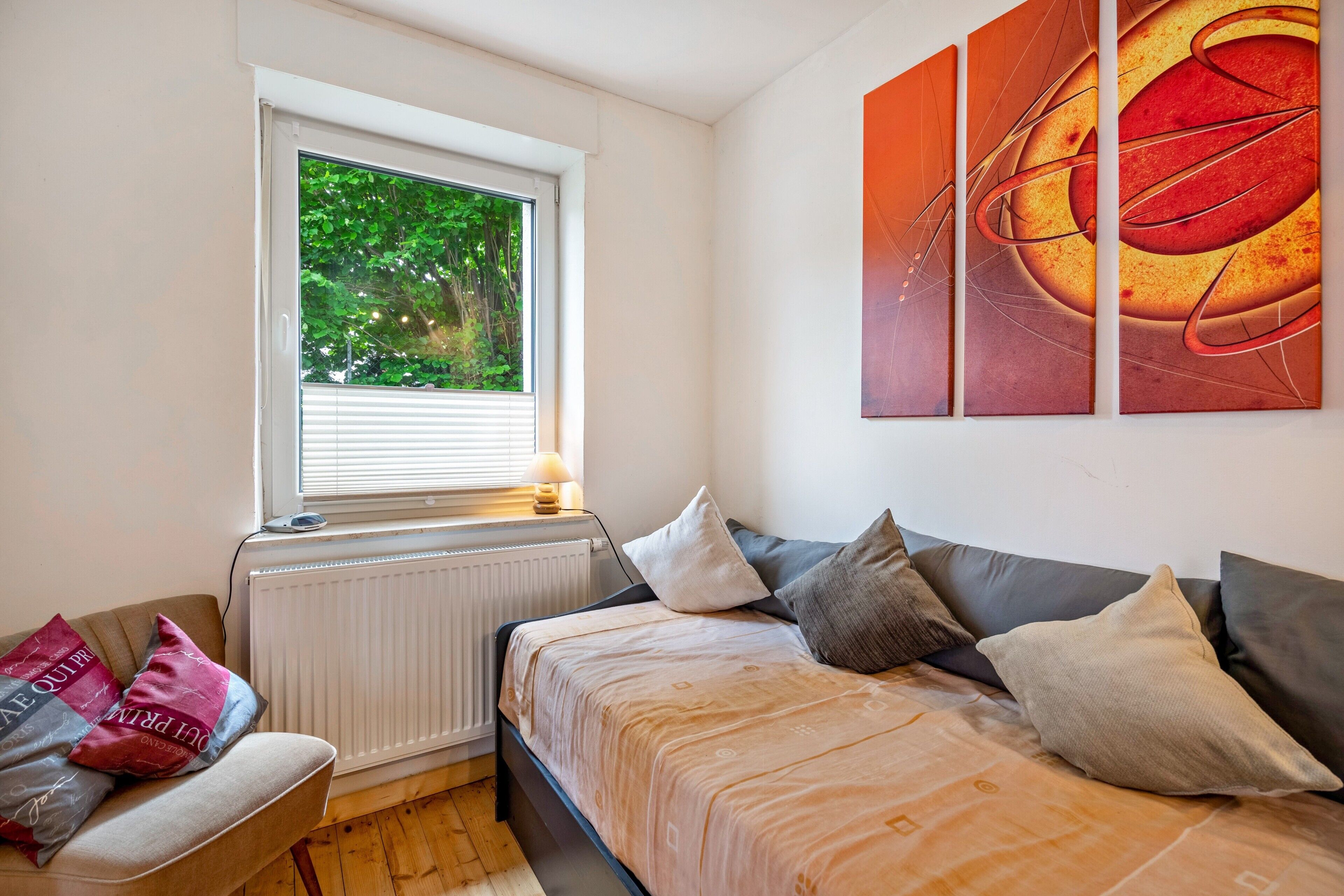 Et soveværelse med seng, vindue med rullegardiner, radiator, sofa og et billede på væggen.