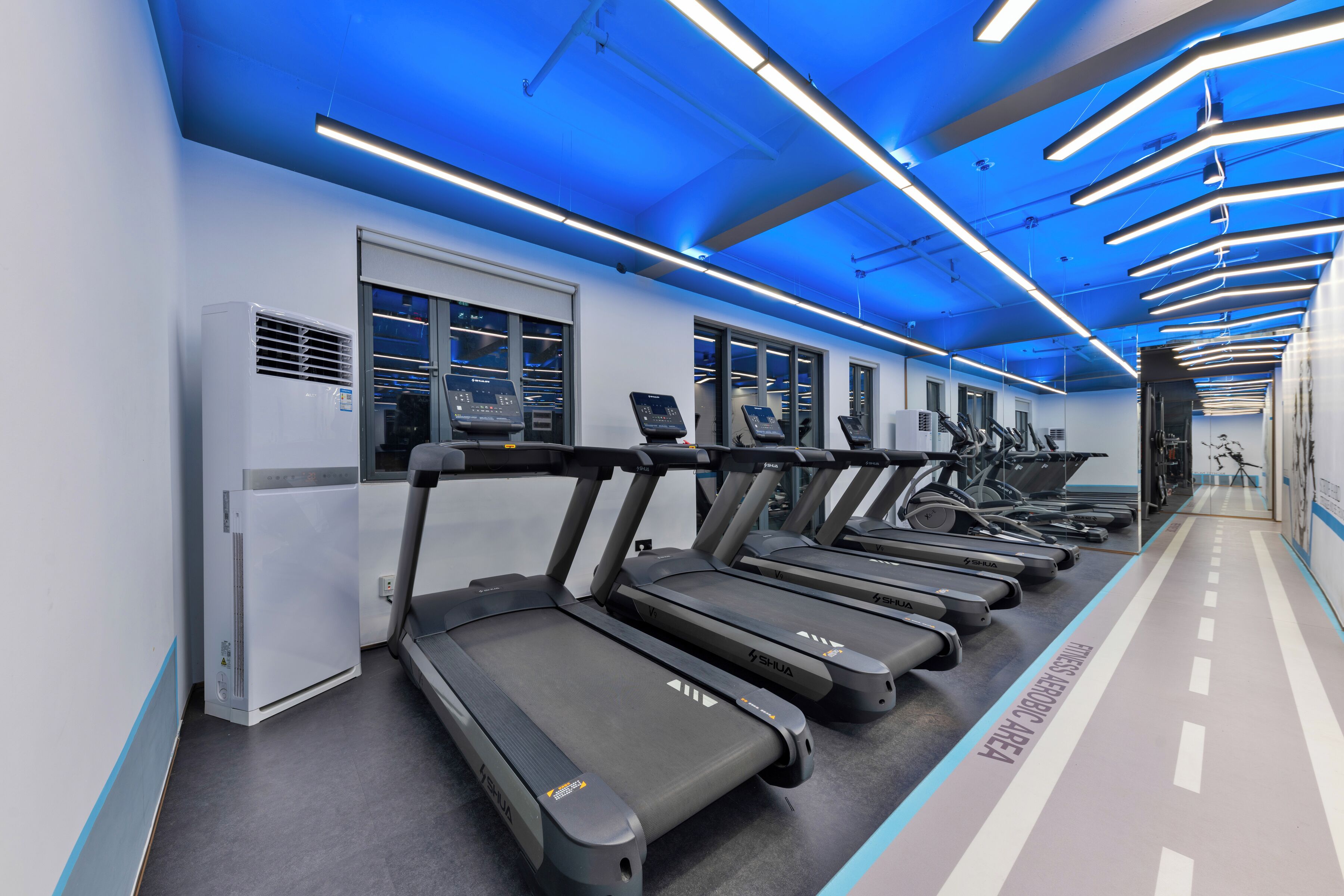 Academia com esteiras, ar condicionado na parede e teto iluminado em azul.