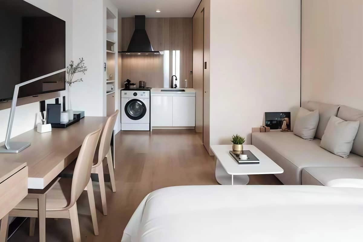 A modern hotel room with a bed, a desk, a sofa, a TV, a washing machine, and a kitchenette.