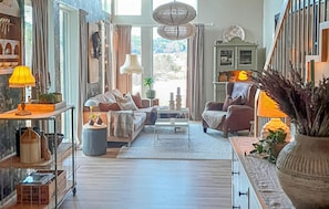 A spacious living area with a sofa, armchairs, a coffee table, and a fireplace. There is a large window providing natural light and a view of the outdoors.