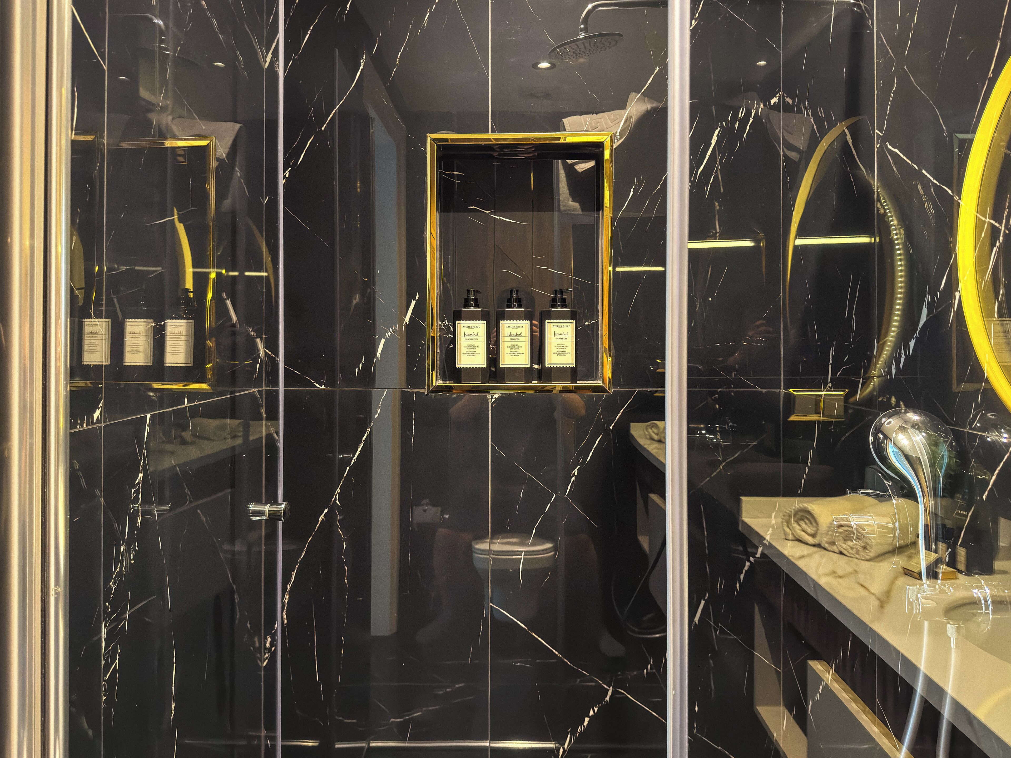 Een badkamer met een wand van zwart marmer, een spiegel met gouden lijst en een plank met toiletartikelen.