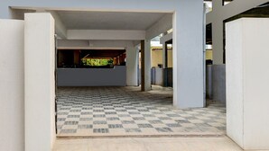 A covered parking area with a checkered floor pattern, concrete pillars, and a view of a building with a green roof. - Deluxe Hotel Nakshatra (Patancheruvu)