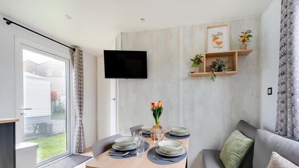 Un salon moderne comprenant un coin repas, un téléviseur à écran plat fixé au mur et une petite étagère pour les plantes.