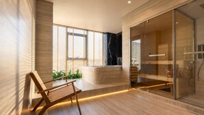 A modern bathroom with a glass shower enclosure, a wooden bench, and a seating area with plants.