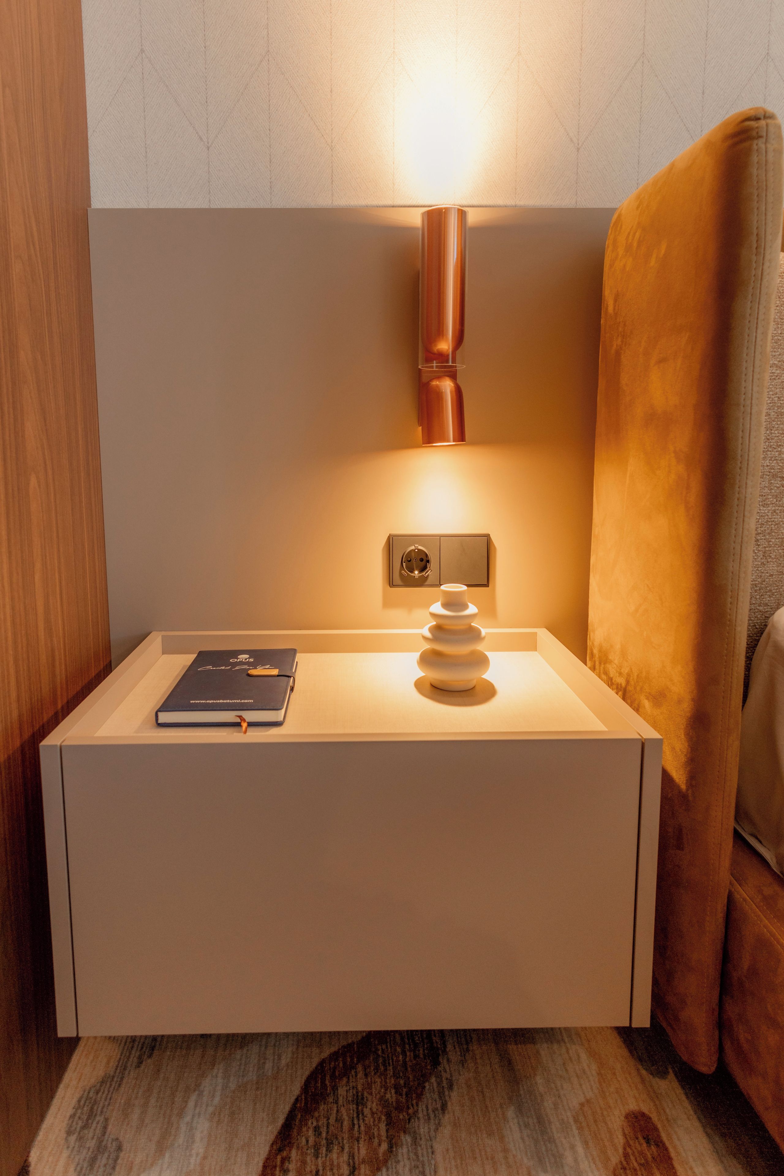 A bedside table with a book, a lamp, and a power outlet.