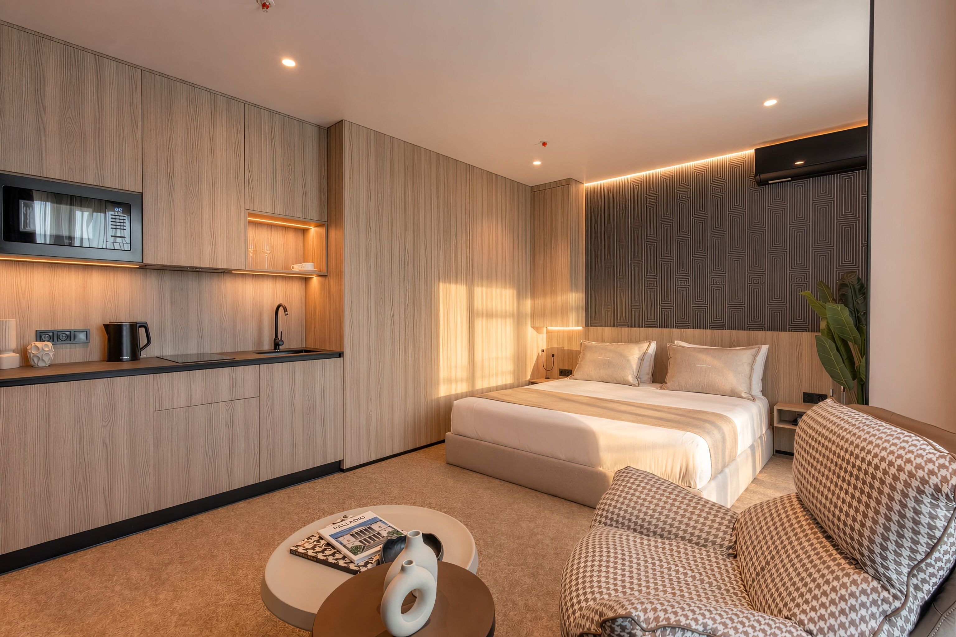Ein modernes Hotelzimmer mit einem großen Bett, einer Sitzecke, einem kleinen Tisch, einer integrierten Küchenzeile und einem an der Wand befestigten Fernseher.
