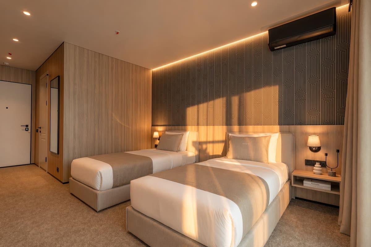 Habitación de hotel moderna con dos camas, un muro decorativo de madera, un papel tapiz estampado y una mesita de noche con lámpara.