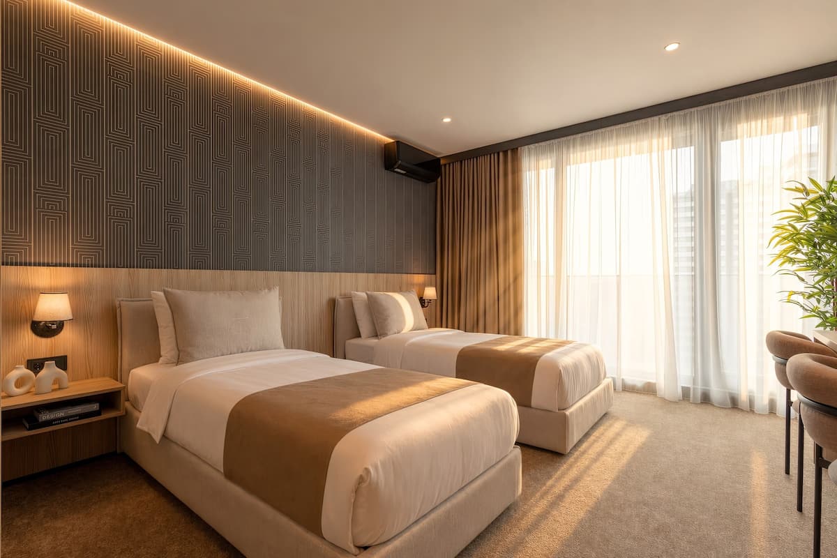 Habitación de hotel con dos camas, un ventanal con cortinas traslúcidas, aire acondicionado montado en la pared y una lámpara de noche.