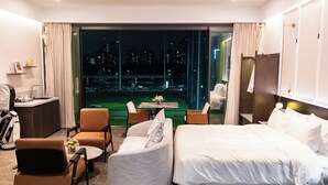 A hotel room with a bed, two chairs, a small table, a desk, and a view of a cityscape at night. - Golf Suite Hotel (Shenzhen)