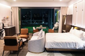 Quarto de hotel com cama, duas cadeiras, uma mesa pequena, escrivaninha e vista para o horizonte urbano à noite.