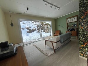 Ein Zimmer mit einer Couch, einem Schreibtisch mit einem Laptop, einem Bücherregal und einem großen Fenster mit Blick auf schneebedeckte Felsen.