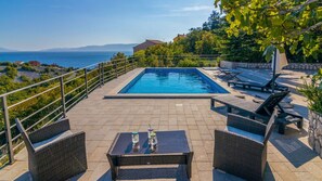 Une piscine entourée d’un mur de pierre et de verdure, avec des chaises longues et une table, offrant une vue sur un paysage côtier.