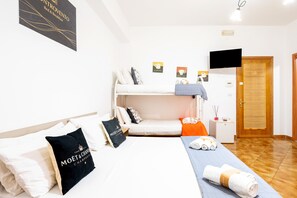 A hotel room with a bunk bed, a bedside table with a lamp, a TV mounted on the wall, and a wooden door. - Quadruple Room with Private Bathroom (Porto Cesareo)