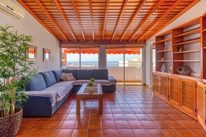 A living room with a wooden ceiling, a blue sofa, a coffee table, and a large window overlooking the sea.