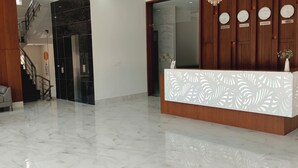 A reception desk with a white decorative panel, a grey sofa, a small table, and a staircase in the background. - Hotel Harsh Vilas (Basai)