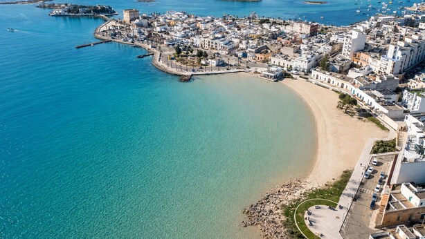Vista aérea de un pueblo costero con playa, aguas azules cristalinas y una mezcla de edificios residenciales y comerciales.