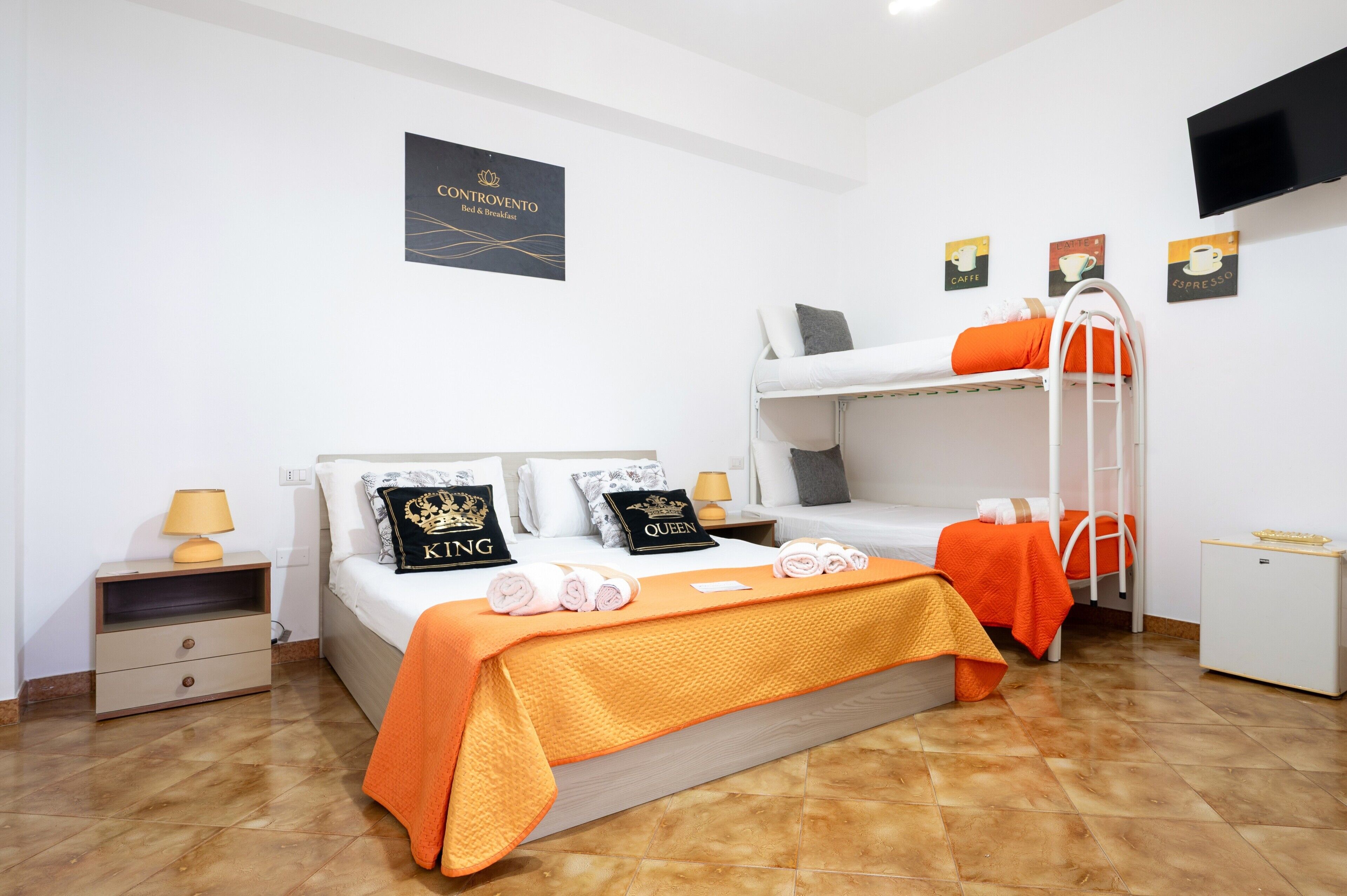 Une chambre d’hôtel avec un lit superposé, un bureau, une chaise, une télévision et un lit recouvert d’une couverture orange.