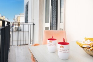 Un balcone con due tazze bianche etichettate 'CONTROVENTO Bed & Breakfast', un cesto in vimini con generi alimentari e vista su edifici sullo sfondo.