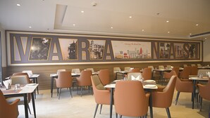 A restaurant with a large wall mural of cityscapes and the word 'NAVA' prominently displayed. Tables are arranged in a spacious layout with orange chairs. - ANANTA KASHI HOTEL (Varanasi)
