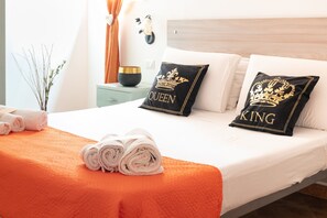 Ein ordentlich bezogenes Bett mit zwei Kissen, auf denen 'QUEEN' und 'KING' stehen, ein aufgerolltes weißes Handtuch und eine kleine Vase mit Zweigen auf einem Nachttisch.
