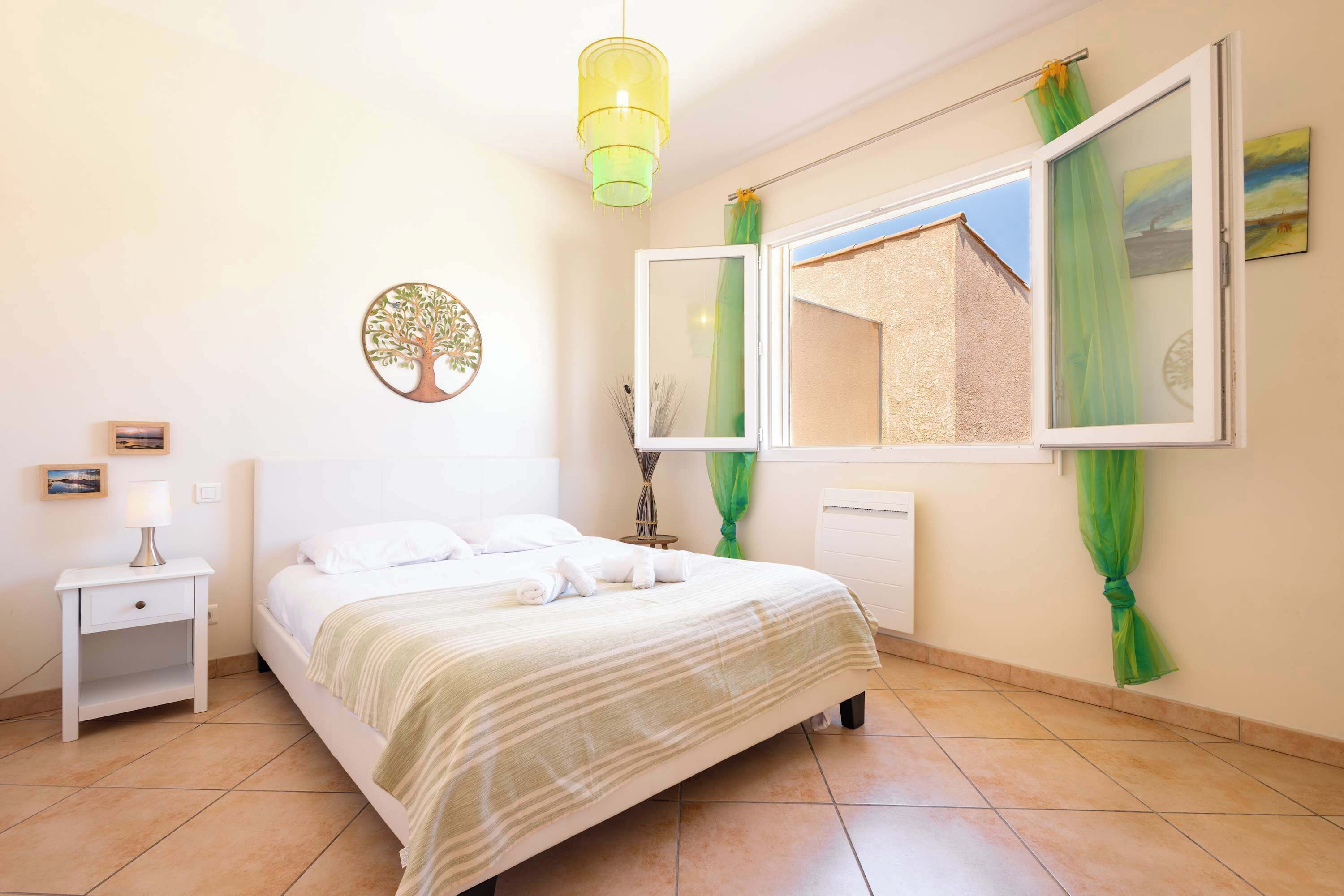 A bedroom with a bed, a nightstand, a lamp, a window with curtains, and a wall-mounted decoration.