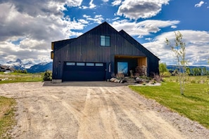 A modern building with a large garage, surrounded by a gravel driveway and grassy areas, with mountains in the background. - Modern Teton View Home in Driggs w/AC (Driggs)
