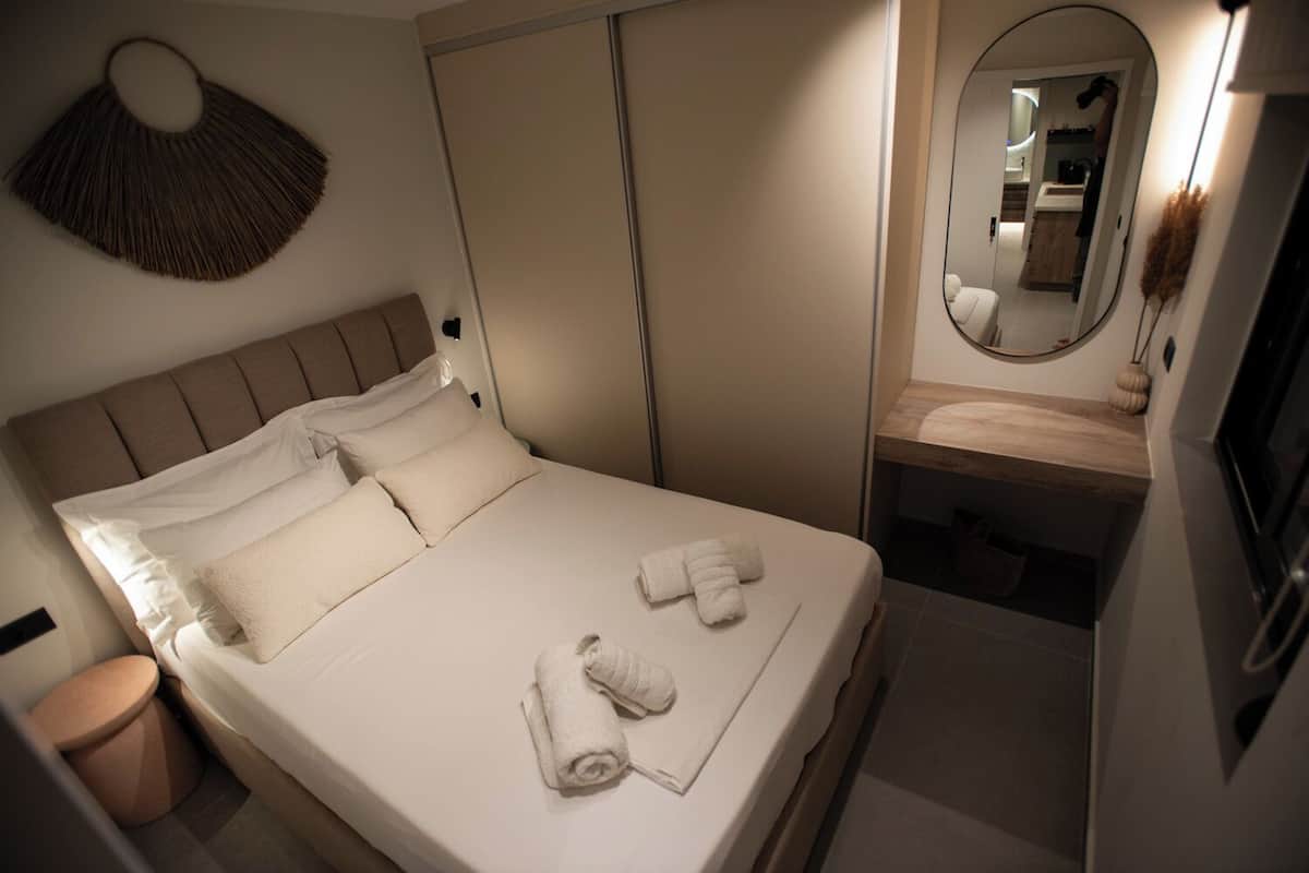 Een net opgemaakt bed met kussens en handdoeken, een nachtkastje, een spiegel en een garderobe.