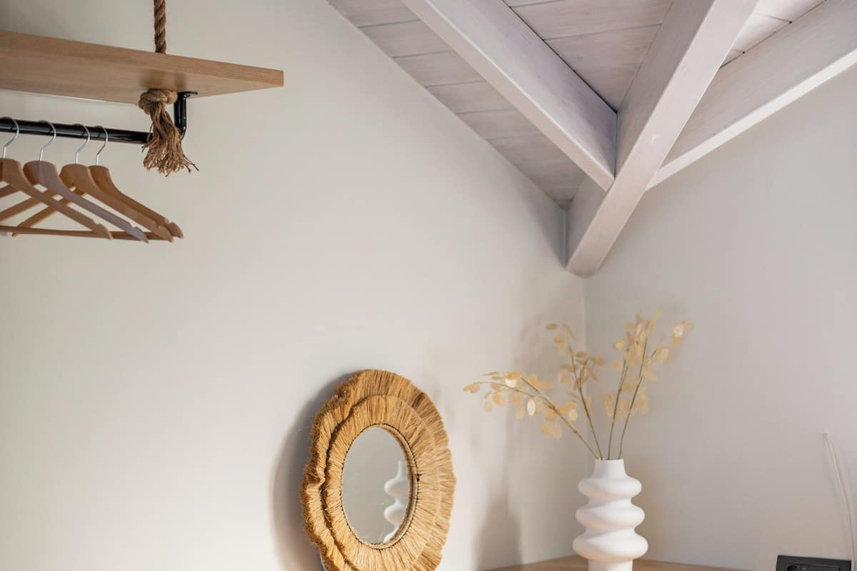 Een slaapkamer met een schuine kap, houten balken, een bed, een nachtkastje, een spiegel, een vaas met gedroogde bloemen en een kapstok met ophangers.