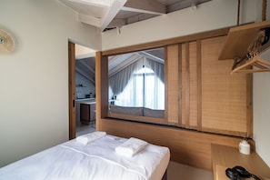 Quarto com cama grande, cabeceira de madeira, mesa de cabeceira com abajur e vista para uma cozinha e varanda através de uma janela.