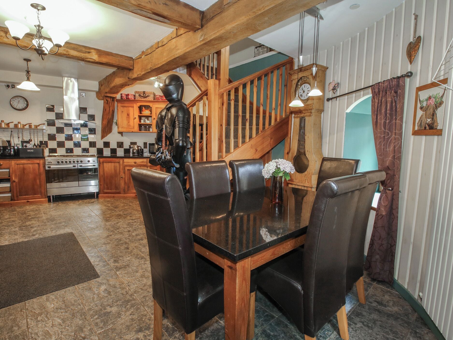 A dining area with a wooden table and leather chairs, a kitchen with checkered tiles, and a staircase with wooden railings.