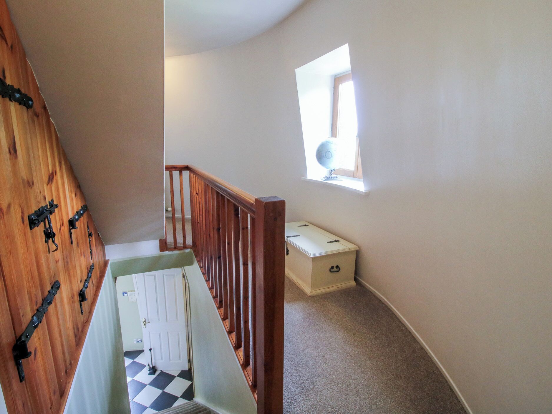 A staircase with a wooden handrail, a checkered floor, a small window with a globe, and a white storage box.