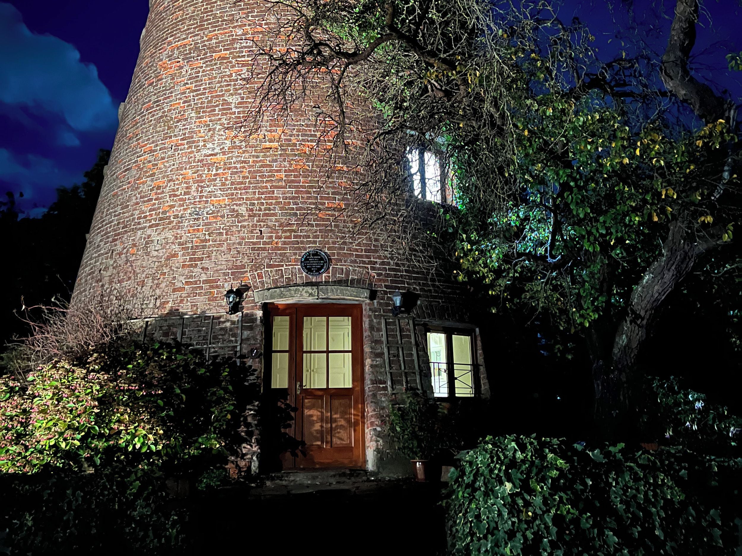 A brick tower with a wooden door and windows, surrounded by greenery.