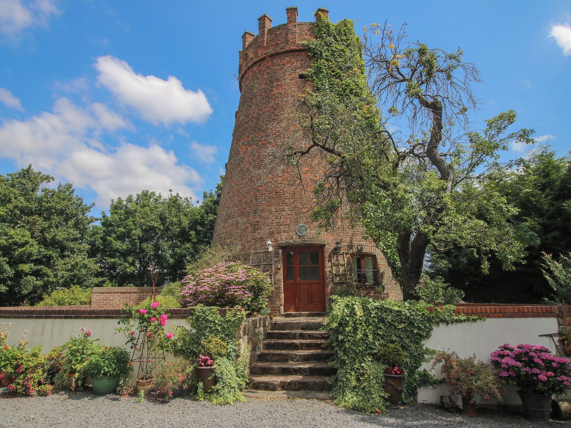 A brick tower with a wooden door and steps leading up to it, surrounded by greenery and flowering plants.