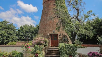 A brick tower with a wooden door and steps leading up to it, surrounded by greenery and flowering plants.