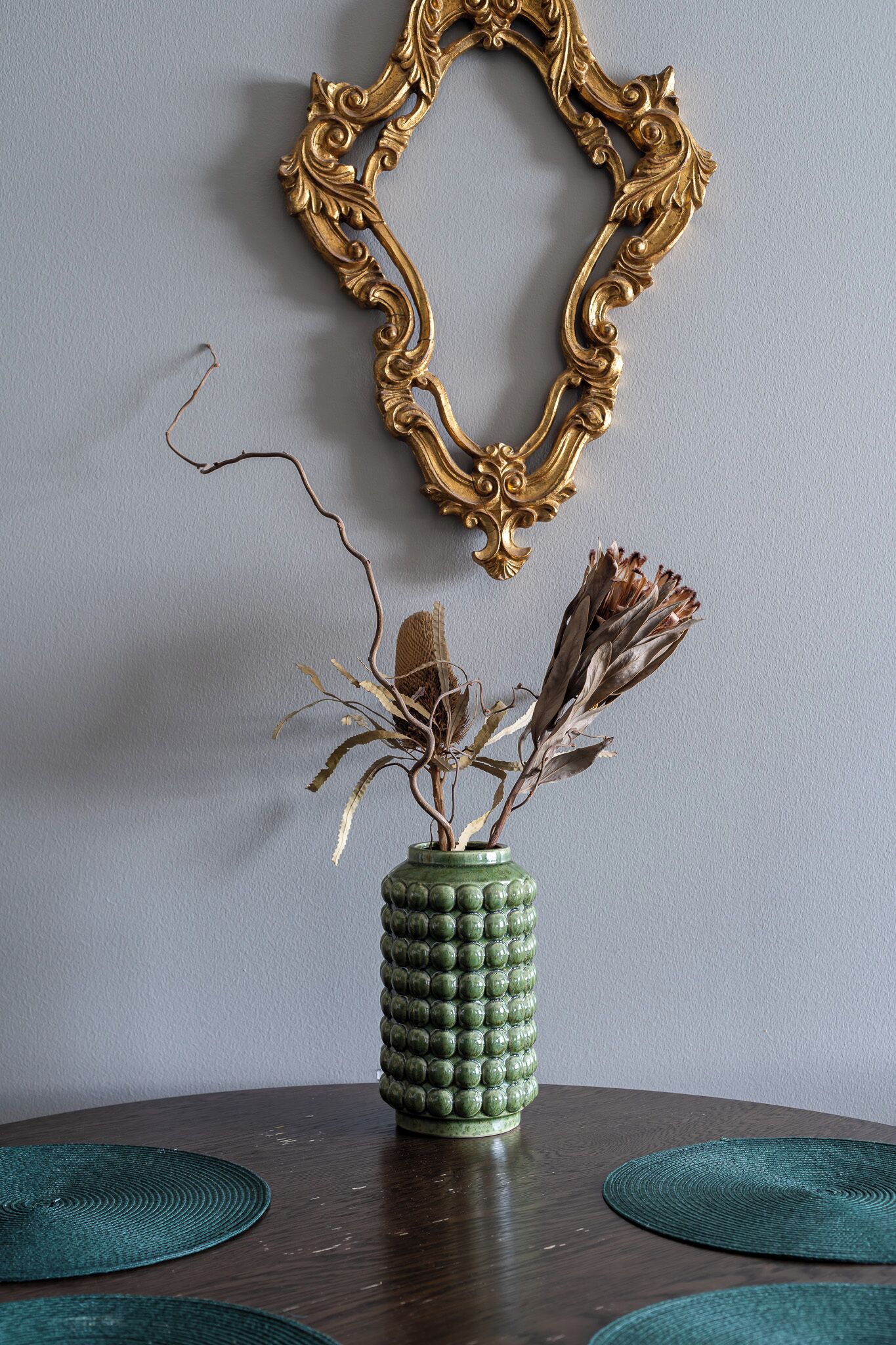 A green vase with dried plants on a wooden table, a gold ornate mirror on the wall, and a textured green placemat.