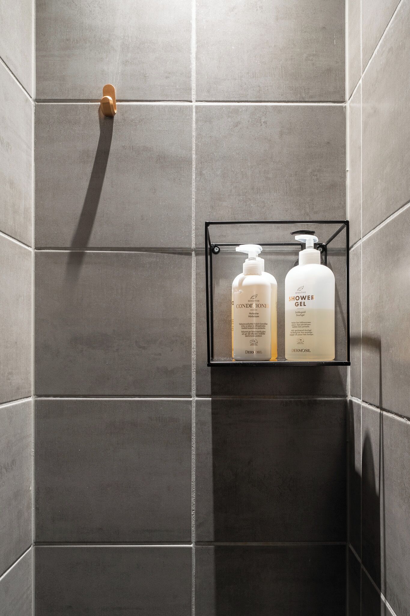 Un baño con baldosas oscuras, cabezal de ducha y repisa con dos botellas de gel de ducha y acondicionador.