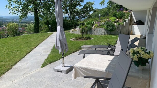An outdoor patio with a table and chairs, a pathway, and a view of a green landscape and distant buildings.