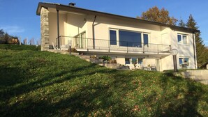 A two-story house with a balcony, a stone facade, and a grassy hill in the foreground.